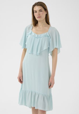 Frau in einem hellblauen, knielangen Kleid mit flatternden Ärmeln, gerafftem Ausschnitt und Rüschensaum steht vor einem schlichten Hintergrund.