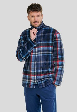 Hombre que lleva una chaqueta de forro polar a cuadros en azul, negro, rojo y blanco, parcialmente abrochada, con una mano en el bolsillo, contra un fondo liso.