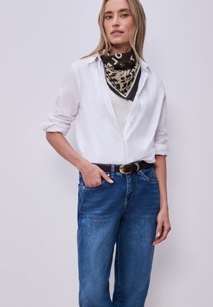 Femme portant une chemise blanche à boutons, un jean bleu avec une ceinture noire, et un foulard léopard autour du cou, debout avec une main dans la poche.
