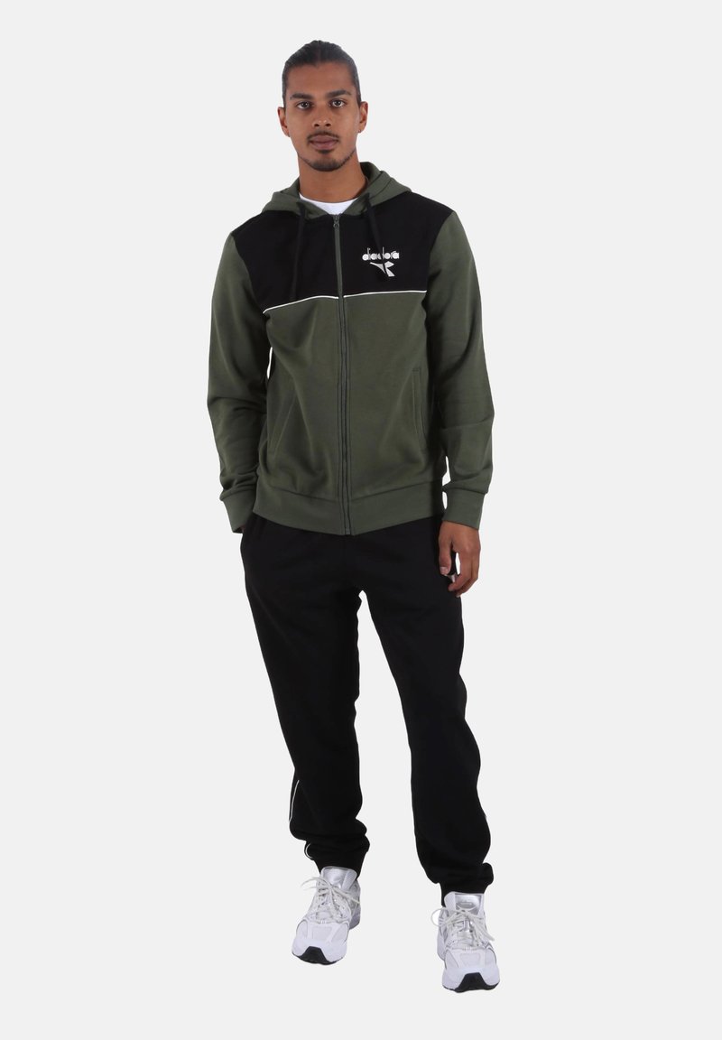 Felpa con cappuccio e cerniera in verde oliva e nero, con accenti bianchi. Indossata con pantaloni jogging neri e scarpe da ginnastica bianche, in piedi con le mani lungo i fianchi.