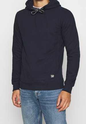 Mężczyzna w granatowej bluzie z kapturem z czarno-białymi troczkami i jasnoniebieskich dżinsach, stojący na jednolitym tle.