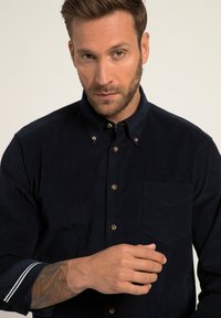 Camicia button-up blu navy con tessuto testurizzato, tasca sul petto e dettagli a righe in contrasto sui polsini. Bottoni in metallo spazzolato.