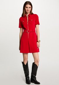 Vestido rojo de manga corta con cremallera frontal, dos bolsillos en el pecho, cintura ceñida y falda plisada. Combinado con botas de cowboy negras hasta la rodilla.