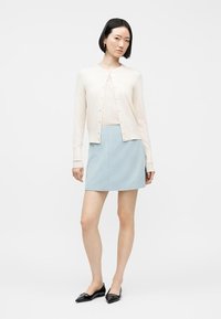 Γυναίκα με κρεμ ζακέτα με κουμπιά, ανοιχτό μπλε μίνι φούστα και μαύρα μυτερά φλατ, όρθια με τα χέρια χαλαρά σε λευκό φόντο.