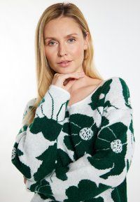 Sweter wykonany na drutach z zielonymi wzorami kwiatowymi na białym tle, z luźnym dekoltem i długimi rękawami. Miękka, teksturowana tkanina.