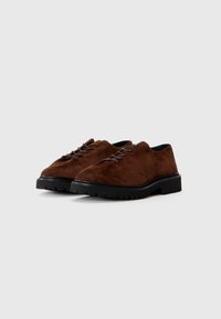 Chaussures en suede marron à lacets avec un bout arrondi et une semelle épaisse en caoutchouc noir présentant un motif de traction. Texture lisse avec des détails de couture discrets.
