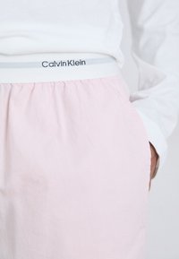 Närbild av en person iklädd en vit långärmad tröja och ljusrosa byxor med en Calvin Klein-resår.