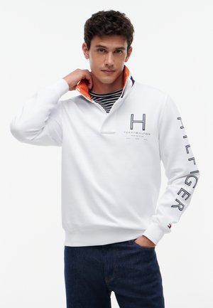 Mand justerer kraven på en hvid Tommy Hilfiger sweatshirt med marineblå og orange foring, marineblå jeans og en stribet skjorte nedenunder.