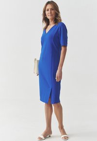 Blaues knielanges Kleid mit V-Ausschnitt und kurzen Ärmeln. Besitzt seitliche Schlitz und eine glatte Textur. Kombination mit weißen Sandalen mit offenem Zehen.