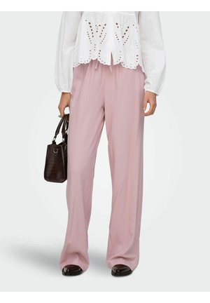 Kobieta w luźnych różowych spodniach i białej bluzce z oczkami, trzyma ciemnobrązową torebkę w lewej ręce, stoi na białym tle.