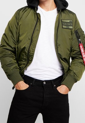 Persoon draagt een opengewerkte olijfgroene bomberjack over een wit shirt en zwarte spijkerbroek, met handen in de zakken en een label aan de mouw van de jas.