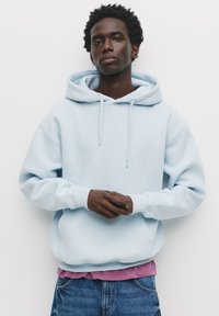Felpa leggera azzurra con coulisse, polsini a coste e vestibilità rilassata, indossata sopra una maglietta viola, abbinata a jeans blu. Design minimalista.