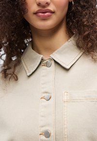 Κοντινό πλάνο ενός ατόμου με σγουρά μαλλιά που φοράει μπεζ πουκάμισο με ασημένια κουμπιά και τσέπη στο στήθος.