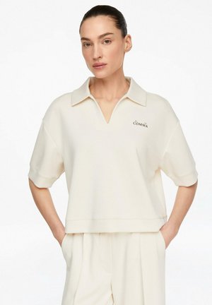 Kobieta o ciemnych włosach zaczesanych do tyłu, ubrana w kremowy krótki rękaw z kołnierzykiem i pasujące spodnie z wysokim stanem, ręce w kieszeniach.