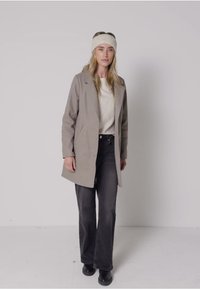 Béžový vlněný kabát s přiléhavým střihem, který má klopový límec a boční kapsy, doplněný černými zvonovými džíny s vysokým pasem a černými botami.