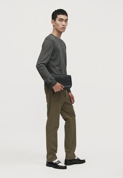 Un homme porte un pull gris foncé, un pantalon olive, des chaussures noires et blanches et une pochette noire devant un fond de studio uni.