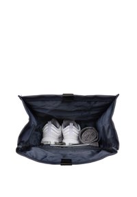 Sac de sport bleu avec une large ouverture, contenant des chaussures de sport blanches et une serviette grise roulée, montrant un intérieur lisse et texturé.