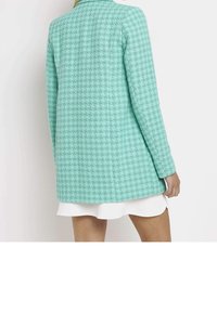 Cappotto verde menta a scacchi con un motivo testurizzato, design a singolo petto, maniche lunghe e taglio dritto, indossato sopra un vestito bianco.