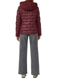 Personne vêtue d'une veste matelassée maroon avec capuche, de jeans gris à jambes larges et de chaussures blanches, se tenant de dos contre un fond blanc.