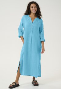Vestito di lino azzurro chiaro con patta di bottoni, maniche a tre quarti, spacchi laterali e tasche. Indossato con sandali neri con dettagli dorati.