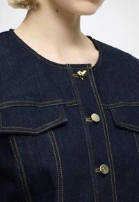 Σκούρα τζιν τζάκετ με κουμπιά σε σχήμα καρδιάς και στρογγυλά σε χρυσό τόνο, δύο τσέπες στο στήθος, διακοσμητικές ραφές και στρογγυλή λαιμόκοψη.