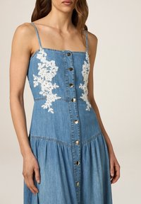 Robe en denim avec des bretelles spaghetti, motif floral blanc brodé sur le bodice, et boutons dorés au centre. Jupe évasée en bas.