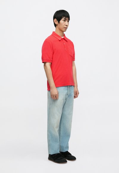 Polo rouge avec une poche, assorti à un jean droit bleu clair. Baskets noires complètent la tenue. Design minimaliste.