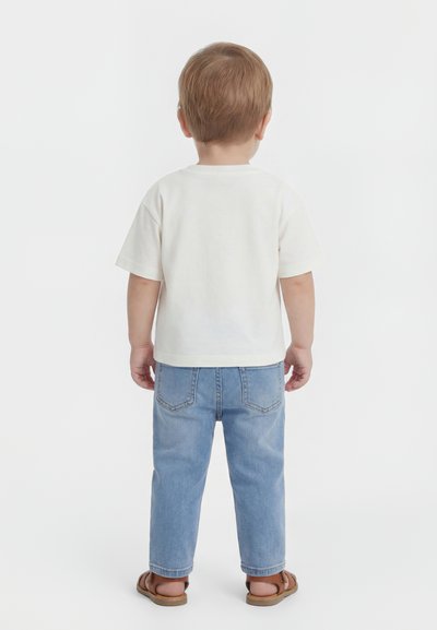 Jeune enfant aux cheveux châtain clair portant un t-shirt blanc à manches courtes, un jean bleu clair et des sandales marron, debout de dos.