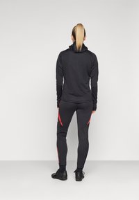 Veste de sport noire et leggings arborant des accents rouges contrastants et une coupe ajustée, fabriqués en tissu extensible avec une texture lisse.