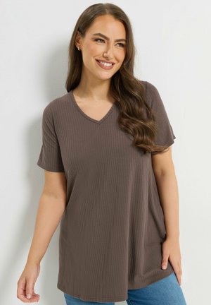 Frau mit langem, welligem Haar lächelt, trägt ein braunes, geripptes Kurzarmshirt mit V-Ausschnitt und blaue Jeans, steht vor einer weißen Wand.