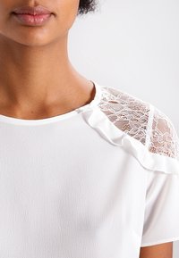 Blouse blanche avec des accents en dentelle transparente sur les épaules, une texture douce, des manches courtes et un détail de col froncé.