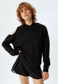 Sudadera negra oversized con diseño de hombros descubiertos, capucha y mangas largas. Hecha de tela suave, combinada con pantalones cortos negros a juego.