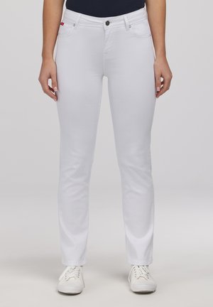 Kobieta ubrana w białe dopasowane jeansy i białe tenisówki na sznurowadła, stojąca na tle jasnego, gładkiego tła.