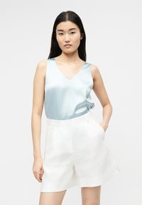 Νεαρή γυναίκα στέκεται με το ένα χέρι στην τσέπη, φορώντας γαλάζιο σατέν αμάνικο τοπ και λευκά σορτς με ψηλή μέση σε ουδέτερο φόντο.