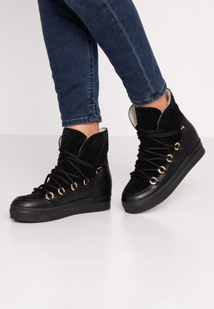 Osoba nosząca czarne wysokie trampki z złotymi przelotkami i czarnymi sznurowadłami, w połączeniu z ciemnoniebieskimi obcisłymi dżinsami.