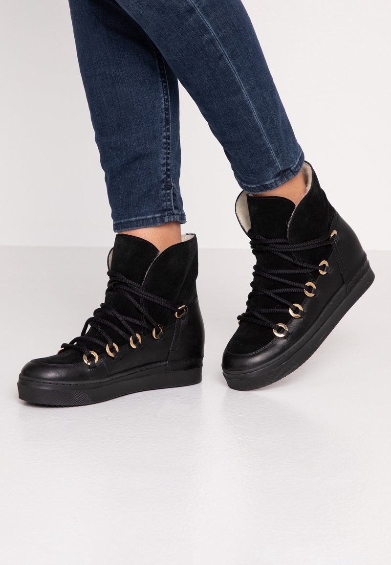 Personne portant des baskets montantes noires avec des œillets dorés et des lacets noirs, associées à un jean skinny bleu foncé.