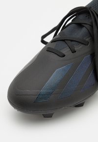 Zwarte synthetische voetbalschoen met een gestructureerd oppervlak, voorzien van donkerblauwe strepen, een verstevigde neus en zwarte veters.