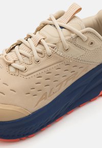 Beige treningssko med teksturert overdel, mesh-detaljer og en blå såle med oransje grepklosser. Inkluderer løkke-snøring og merkevaredetalj.
