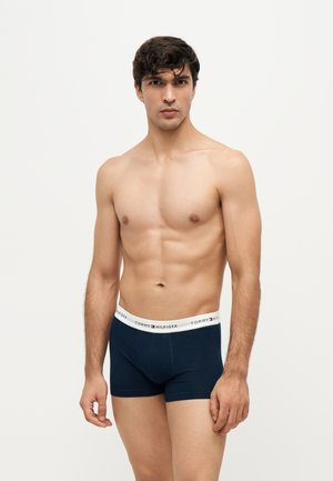 Homme torse nu portant un boxer bleu marine Tommy Hilfiger avec une ceinture blanche à logo, debout devant un fond blanc uni.