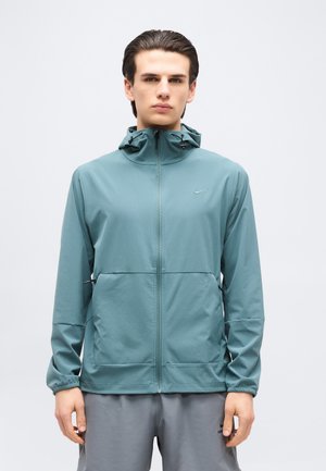 Jeune homme portant une veste à capuche zippée bleu sarcelle et un short Nike gris, debout devant un fond uni gris clair.