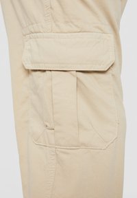 Pierna de pantalones cargo de tela beige que muestra un gran bolsillo lateral rectangular con cierre de solapa y detalles visibles de costuras.