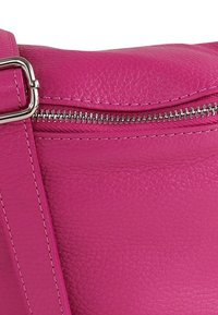 Magenta leren handtas met een gladde textuur, voorzien van een zilveren ritssluiting en verstelbare riem. Zichtbare stiksels voegen details toe.