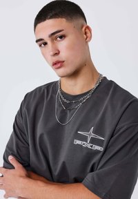 Camiseta oversize gris oscuro con mangas cortas y un logo blanco "PXP", cadenas plateadas superpuestas alrededor del cuello, textura suave.
