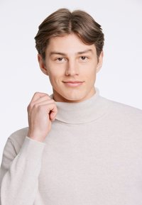 Maglione a collo alto beige chiaro realizzato in materiale morbido, caratterizzato da dettagli a costine sul colletto e sui polsini, indossato da un giovane.