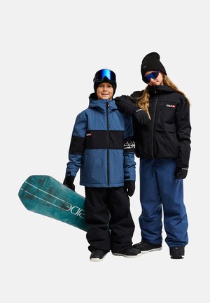 Deux enfants habillés en tenue de snowboard d'hiver, l'un tenant un snowboard, tous deux portant des gants, des vestes et des lunettes.