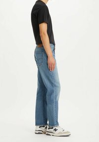 Vaqueros rectos azules con un lavado claro, combinados con una camiseta negra y zapatillas blancas. Presenta cinco bolsillos y un parche de cuero marrón.