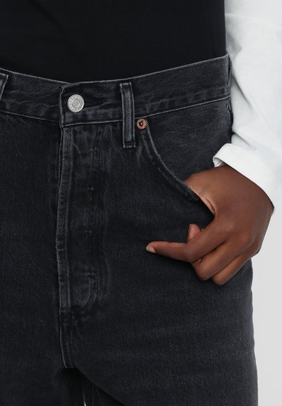 Black denim jeans with a high waist, featuring a silver button, copper rivet, and a soft texture. Hand resting in the front pocket.