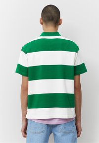 Henkilö, joka on pukeutunut vihreä-valkoiseen leveäraitavaan lyhythihaiseen paitaan, lila tee-paitaan ja vaaleansinisiin farkkuihin, seisoo selkä kameraan päin.