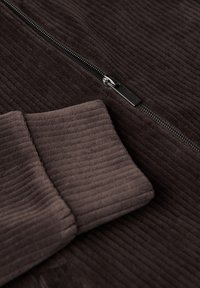 Brun kordflöjlsjacka med en ribbad textur, utrustad med en silverfärgad dragkedja och ribbade muddar. Ytan är mjuk och sammetsliknande.