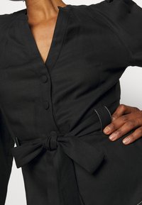 Musta kietaisumekko, jossa on syvä V-kaula-aukko, nappisuljenta ja sidottava vyö. Sileä kangas, jossa on kontrastiväriommelta vyössä.